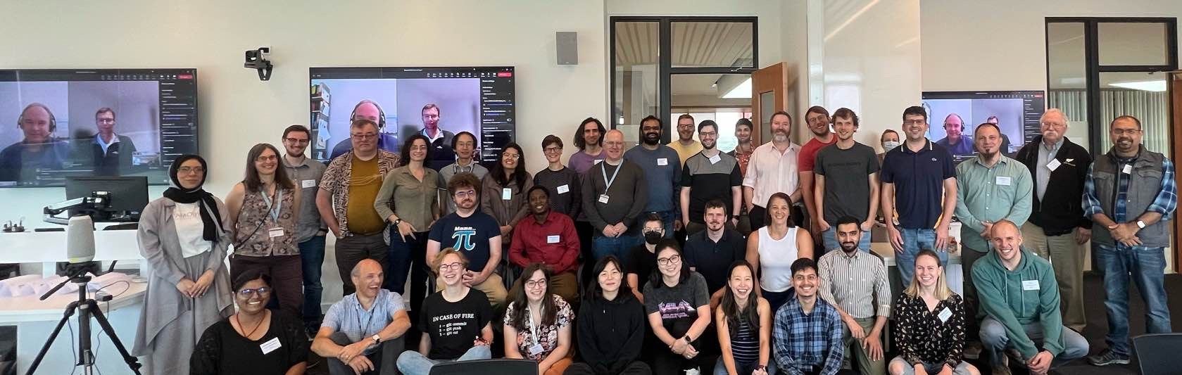 38 participants in-person in a classroom space: 1st row sitting on floor, 2nd row kneeling or sitting in chairs, 3rd row standing. Two remote participants shown on video conference in screen to the left of the group.