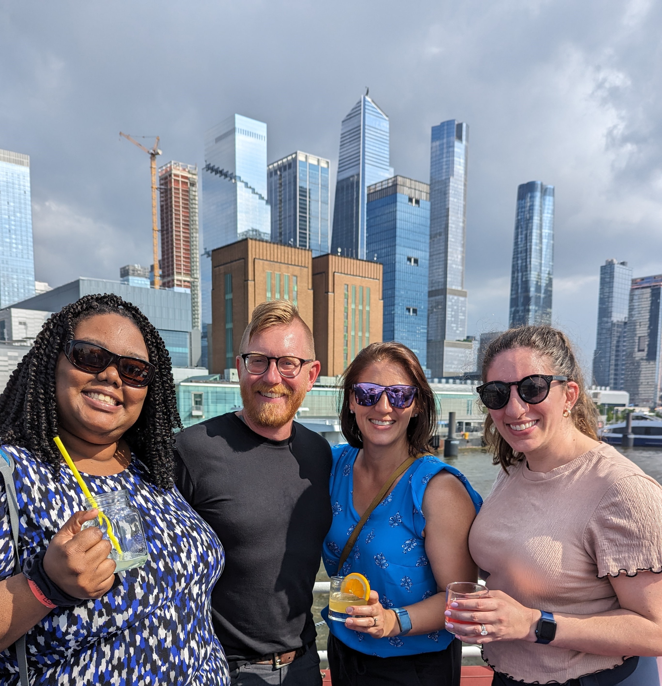 Meghan, Shannon,and Victoria are wearing sunglasses and holding cocktails. Photo taken on a boat; background is water and shiny, tall city buildings.