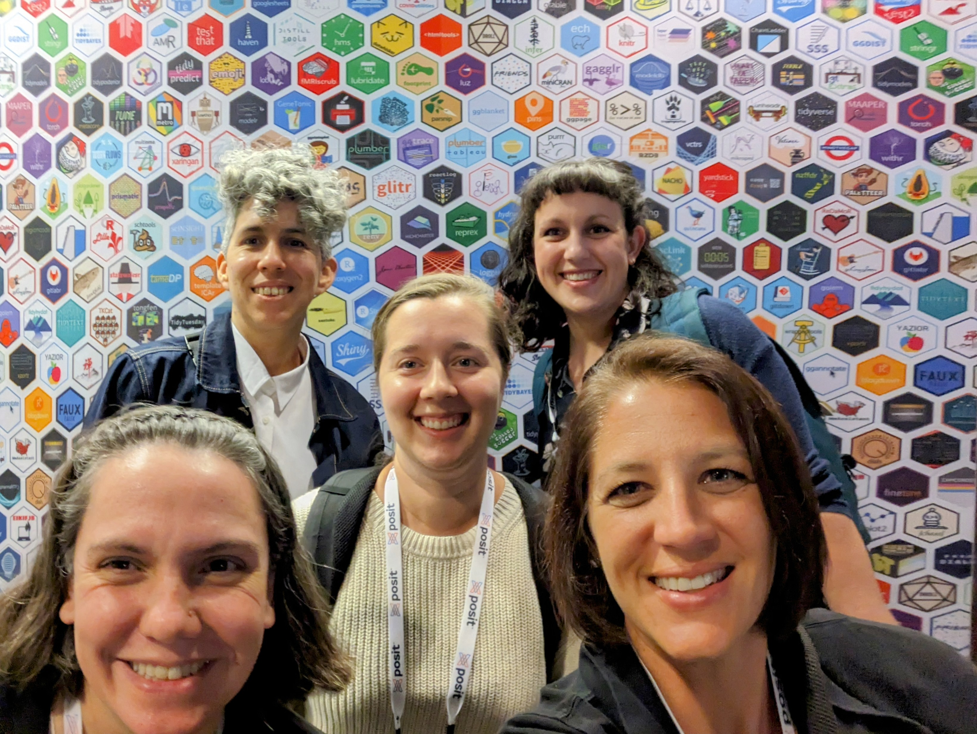 5 women standing together taking a selfie in front of a wall full of hex stickers.