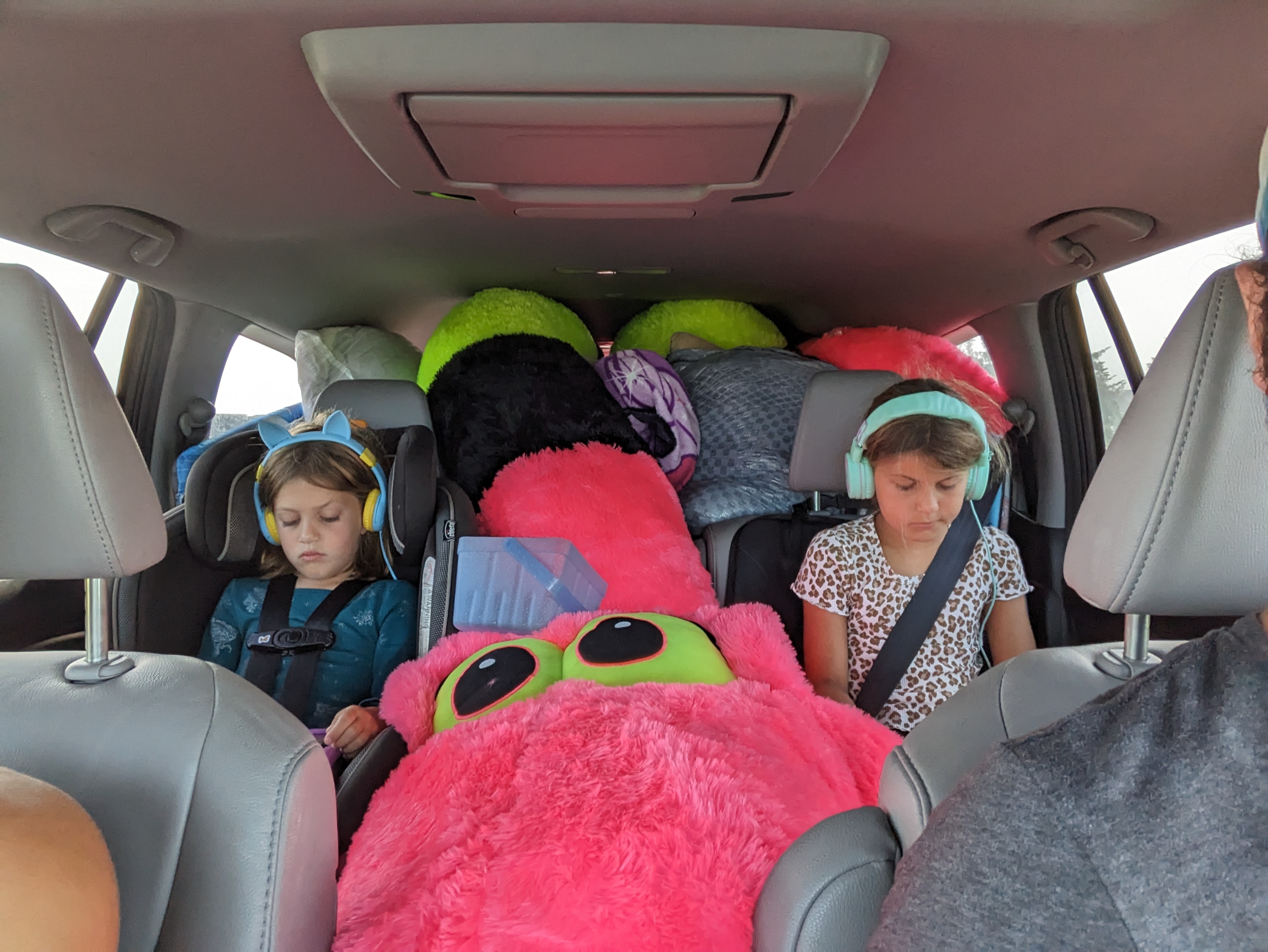 Photo taken from the front seat of a car looking back; two girls are wearing head phones and looking down at ipads. In between is a giant pink stuffed snake with bright yellow eyes.